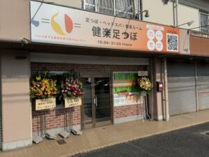 健楽足つぼの店舗外観。看板には営業時間やサービス内容が記載され、入口には開店祝いの花が並んでいる。